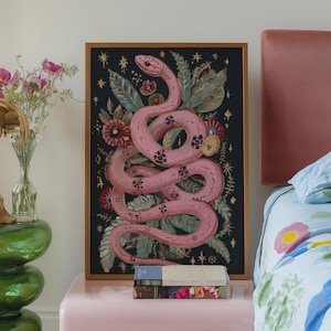 May include: A pink snake with black spots is curled around a floral design on a black background. The artwork is framed in gold and hangs above a pink bedside table with a book on top.