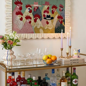 K&ouml;nnte beinhalten: Ein goldener Servierwagen mit Glasplatte, auf dem verschiedene Spirituosenflaschen, Gl&auml;ser und eine Blumenvase stehen. Dar&uuml;ber h&auml;ngt ein gerahmtes Kunstwerk mit H&uuml;hnern, die H&uuml;te tragen. Kerzen und B&uuml;cher sind ebenfalls zu sehen.