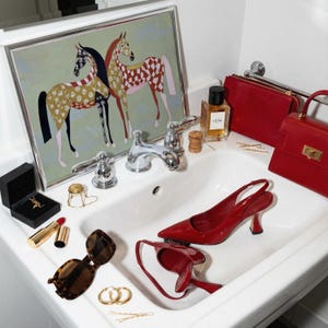 May include: A collection of fashion accessories and beauty products arranged in a bathroom sink. Items include red high heels, handbags, sunglasses, jewelry, and perfume. A framed artwork of two horses hangs above the sink.