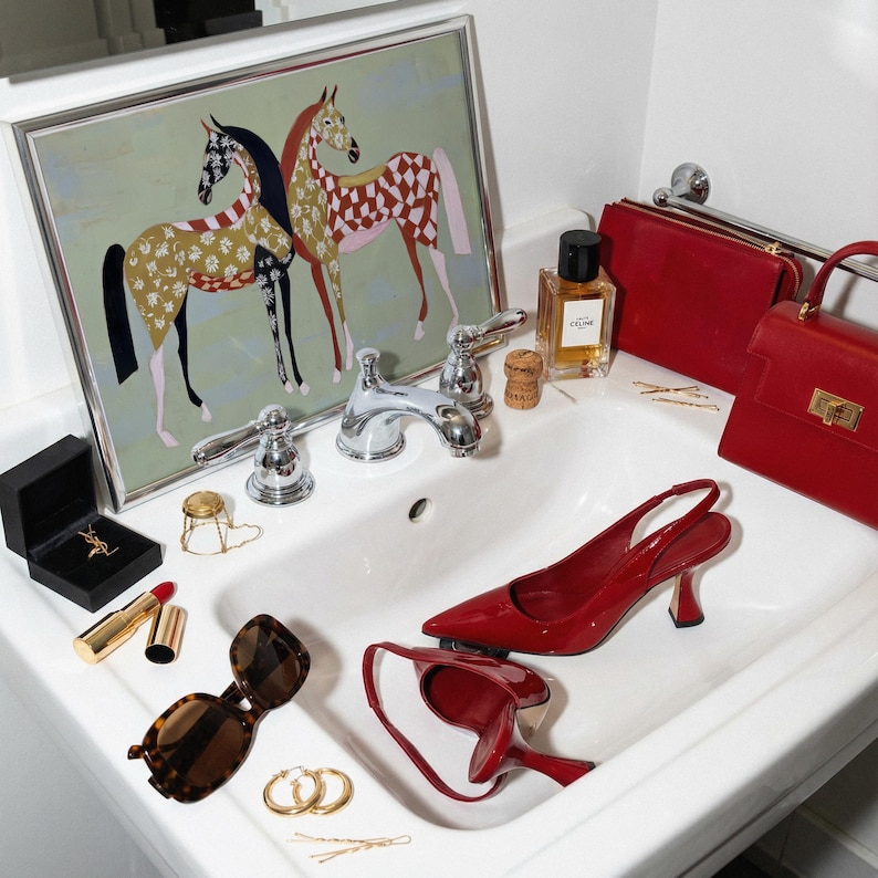May include: A collection of fashion accessories and beauty products arranged in a bathroom sink. Items include red high heels, handbags, sunglasses, jewelry, and perfume. A framed artwork of two horses hangs above the sink.