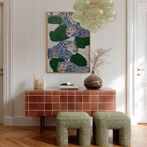 May include: An interior scene featuring a framed artwork with lion illustrations in blue and white, above a brown tiled cabinet. Two patterned stools with a green and black design sit in front of the cabinet. A glass chandelier and a vase with dried branches complete the decor.