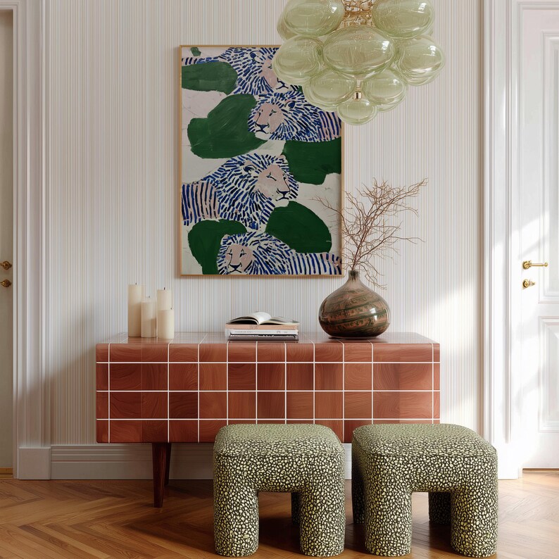 May include: An interior scene featuring a framed artwork with lion illustrations in blue and white, above a brown tiled cabinet. Two patterned stools with a green and black design sit in front of the cabinet. A glass chandelier and a vase with dried branches complete the decor.