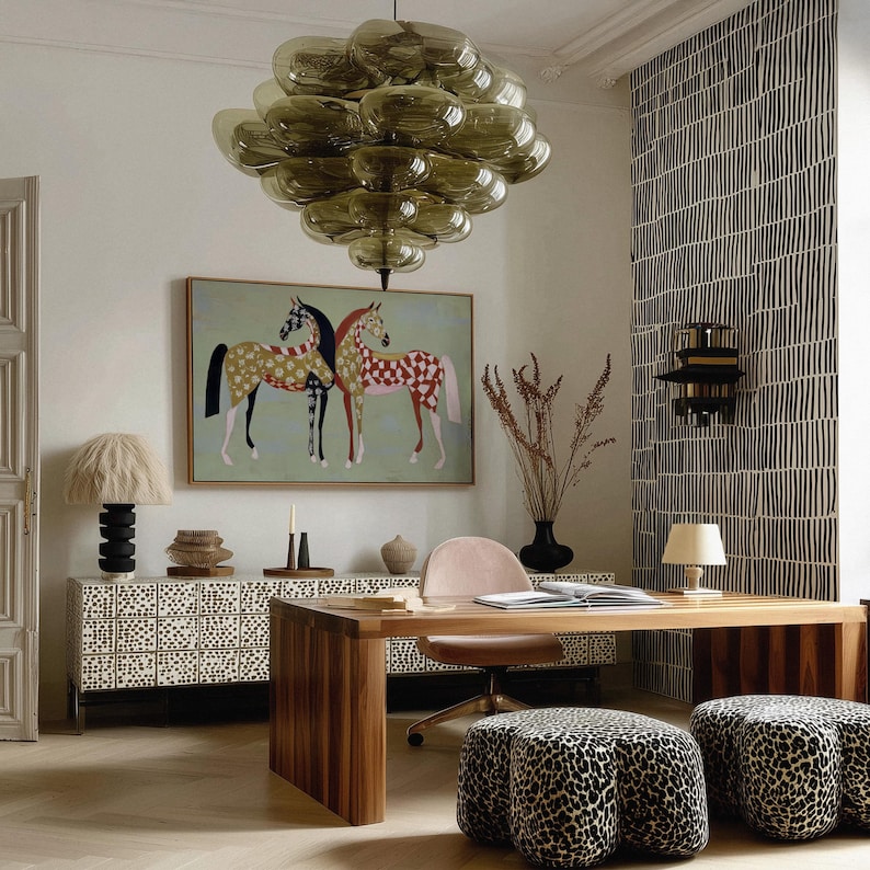 May include: An interior design scene featuring a large, olive-green glass chandelier, a framed painting of two horses, and a wooden desk. The room includes a patterned cabinet, leopard-print ottomans, and a black and white patterned wall.