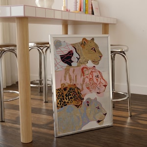 May include: A rectangular silver-framed wall art print featuring a vertical stack of four stylised big cat heads. The illustration includes a white and black patterned lion, a tan lion, a pink tiger, and a blue leopard, all rendered in a detailed line art style.