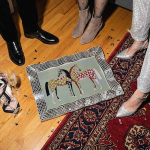May include: A rectangular decorative tray with a mosaic mirrored frame. The tray features two stylized horses with patterned coats on a light green background. The scene is set on a wooden floor with a red rug.
