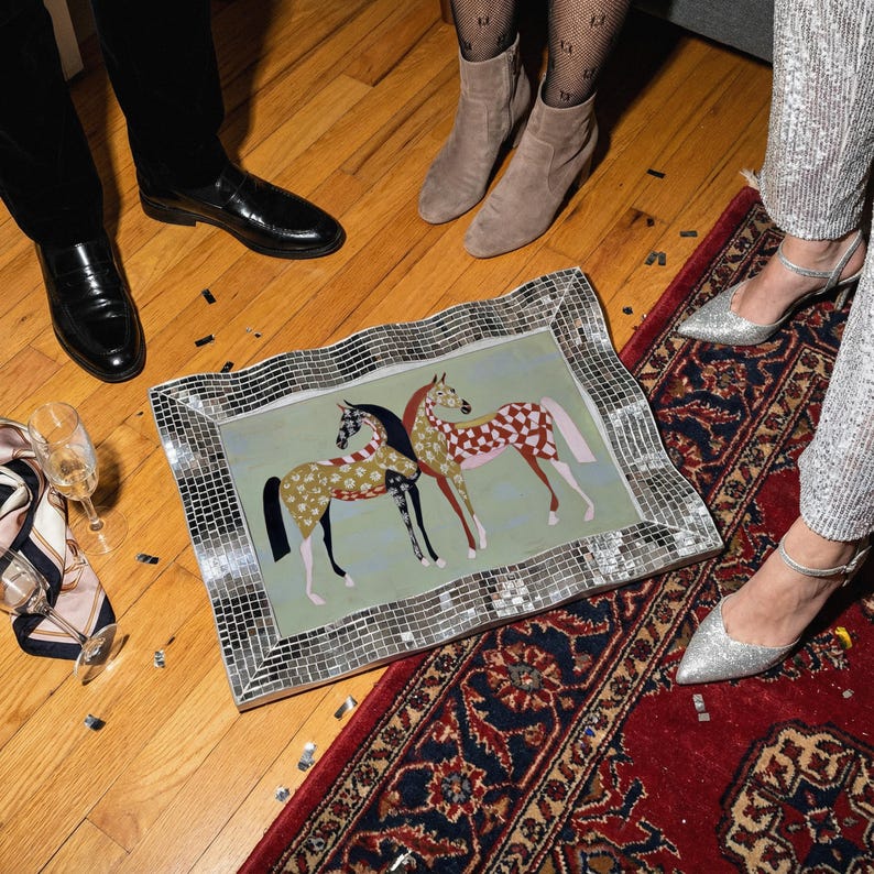 May include: A rectangular decorative tray with a mosaic mirrored frame. The tray features two stylized horses with patterned coats on a light green background. The scene is set on a wooden floor with a red rug.