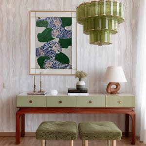 May include: A stylish interior scene featuring a green and wood console table with drawers, topped with decor. Above, a framed artwork depicts lions. Two green patterned stools sit below. A green glass chandelier hangs from the ceiling.