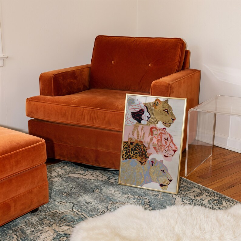 May include: A rectangular framed print featuring a vertical stack of four stylised big cat heads in shades of gold, pink, and blue, resting against a burnt orange velvet armchair with two tufted buttons on the back cushion.