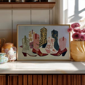 May include: A framed artwork featuring a collection of colorful cowboy boots. The boots showcase various patterns, including floral, leopard print, and textured designs. The artwork is displayed on a white countertop with a wooden cabinet in the background.