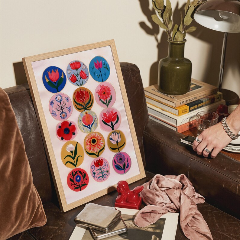 May include: A framed art print featuring colorful floral designs in circular patterns. The artwork is displayed in a light wood frame and rests on a brown leather couch. A red sculpture and a stack of books are also visible.