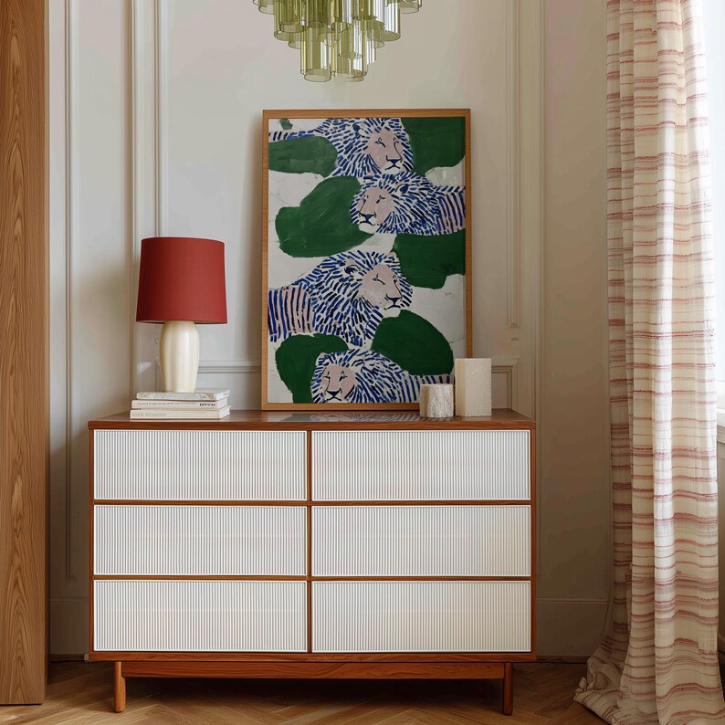 May include: A wooden dresser with six white ribbed drawers and a brown frame. A framed artwork of lions with blue and white manes and green foliage hangs above. A red lamp and books sit on top of the dresser. A green chandelier hangs from the ceiling.