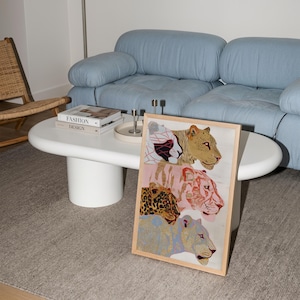 May include: A rectangular framed wall art print featuring four stacked stylised big cats. The top cat is white with black and red geometric patterns, followed by a golden-brown cat, a pink tiger, and a blue leopard with gold spots.
