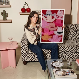 May include: A framed art print featuring a collection of cowboy hats in various shades of pink, orange, and white against a red background. The artwork is held by a person sitting in a zebra-print chair. The room has a pink table and a glass coffee table.