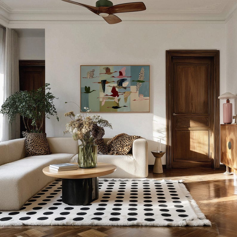 May include: A living room featuring a cream-colored sectional sofa with leopard print pillows, a round wooden coffee table with a black base, and a white area rug with black polka dots. A framed painting of flying birds hangs on the white wall.