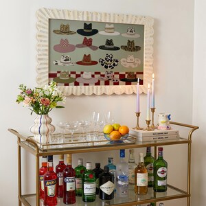 May include: A gold bar cart with various bottles of liquor, glassware, and a vase of flowers. A framed artwork of hats hangs above the cart. Lit candles and books are on the top shelf. The artwork features a variety of hats.