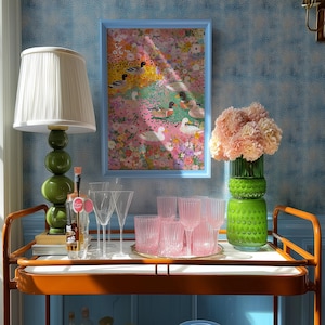 May include: A drinks trolley with a framed picture, a green lamp, and glassware. The artwork depicts ducks in a floral scene. Pink glassware and a green vase with flowers are also on display. The trolley has an orange frame.