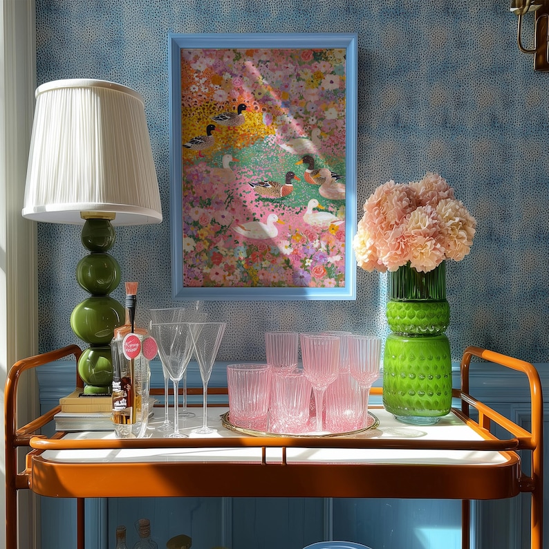 May include: A drinks trolley with a framed picture, a green lamp, and glassware. The artwork depicts ducks in a floral scene. Pink glassware and a green vase with flowers are also on display. The trolley has an orange frame.