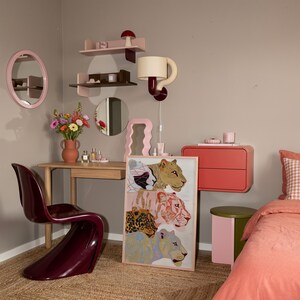 May include: A bedroom interior featuring a wooden desk, a dark burgundy S-shaped chair, and a framed print of four big cats. The room includes pink and brown wall shelves, a wall-mounted lamp, a round mirror, and a coral-coloured floating drawer unit.