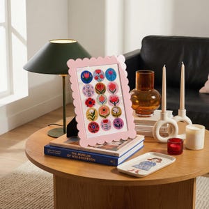 May include: A pink scalloped-edge picture frame displaying colorful floral art. A dark green lamp, a brown glass vase, white candle holders, and a small tray with a girl illustration are on a wooden table. Books are stacked below the frame.