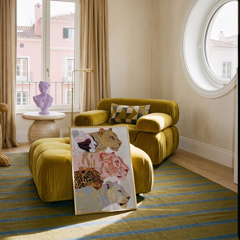 May include: A rectangular framed art print featuring four stacked stylised big cat heads in shades of tan, pink, brown, and light blue, resting against a mustard yellow tufted armchair and ottoman set with a geometric patterned throw pillow.