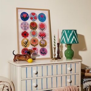 May include: A framed floral art print with colorful flower designs, a green lamp with a patterned shade, and a ceramic dachshund figurine on a striped dresser. The dresser has blue stripes and white knobs.