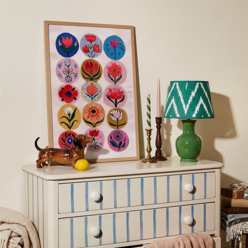 May include: A framed floral art print with colorful flower designs, a green lamp with a patterned shade, and a ceramic dachshund figurine on a striped dresser. The dresser has blue stripes and white knobs.