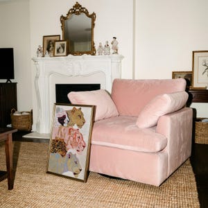 May include: A rectangular framed print featuring a vertical stack of four stylised big cat illustrations in shades of gold, pink, brown, and blue, positioned next to a plush, light pink velvet armchair with a deep seat and two matching cushions.