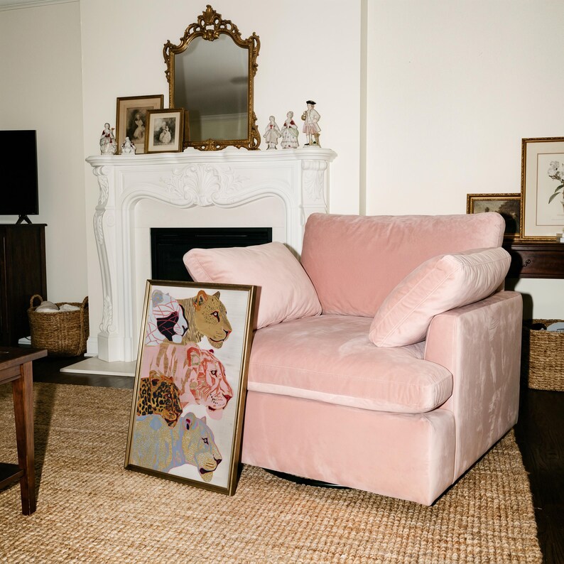 May include: A rectangular framed print featuring a vertical stack of four stylised big cat illustrations in shades of gold, pink, brown, and blue, positioned next to a plush, light pink velvet armchair with a deep seat and two matching cushions.