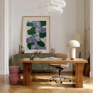 May include: An interior design scene featuring a wooden desk, a patterned cabinet, and a framed artwork with lion illustrations. A desk lamp, a chair, and a decorative stool are also present. The room is illuminated by natural light and a modern chandelier.