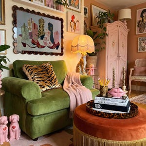 May include: A cozy living room scene with a green velvet armchair, a patterned pillow, and a pink throw blanket. A framed artwork of cowboy boots hangs above the chair. An orange ottoman with a fringed base and a decorative tray is in front of the chair.