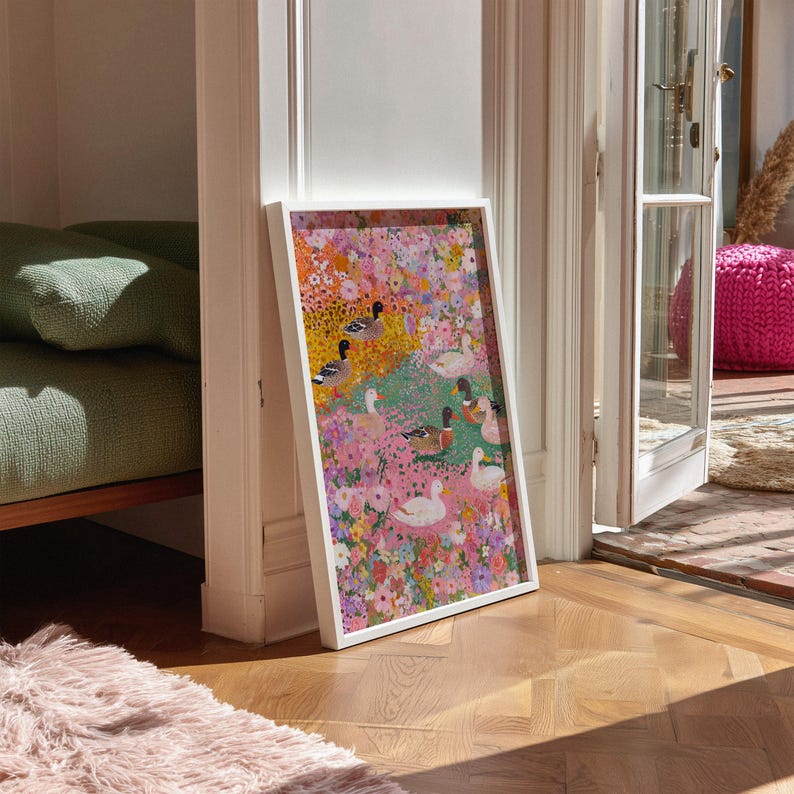 May include: A framed art print depicting ducks in a floral landscape. The print, in a white frame, leans against a white door frame. The artwork's colours include pink, yellow, green, and blue, creating a vibrant and cheerful aesthetic.