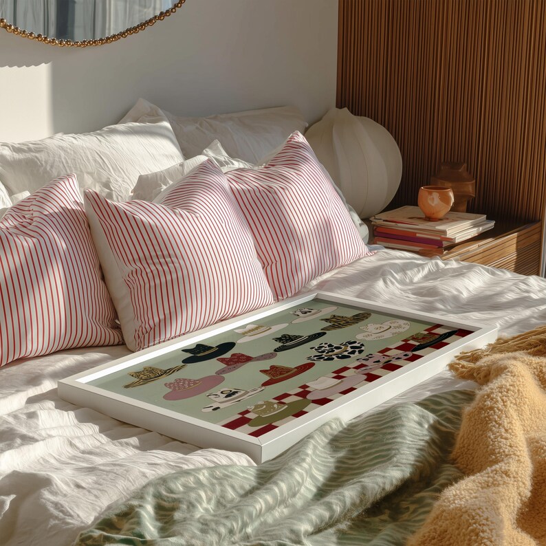 May include: A white framed art print featuring a collection of hats in various styles and colors on a light green background. The print is displayed on a bed with red and white striped pillows and a white comforter. A gold-framed mirror is visible.