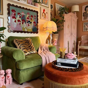 K&ouml;nnte beinhalten: Ein gem&uuml;tliches Wohnzimmer mit einem gr&uuml;nen Samtsessel, einer rosa Decke und einem runden Hocker. Ein dekoratives Gem&auml;lde von H&uuml;hnern h&auml;ngt &uuml;ber dem Stuhl. Eine Leopardenmuster-Lampe und ein rosa Schrank erg&auml;nzen die Einrichtung.