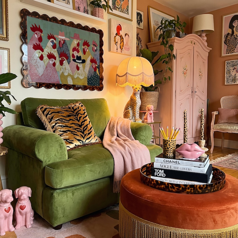K&ouml;nnte beinhalten: Ein gem&uuml;tliches Wohnzimmer mit einem gr&uuml;nen Samtsessel, einer rosa Decke und einem runden Hocker. Ein dekoratives Gem&auml;lde von H&uuml;hnern h&auml;ngt &uuml;ber dem Stuhl. Eine Leopardenmuster-Lampe und ein rosa Schrank erg&auml;nzen die Einrichtung.