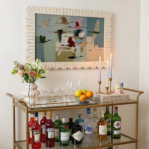 May include: A gold metal bar cart with two glass shelves holding various liquor bottles, glassware, a bowl of citrus, and three taper candles. A scalloped frame surrounds a print of flying birds, and a vase with flowers sits on top.