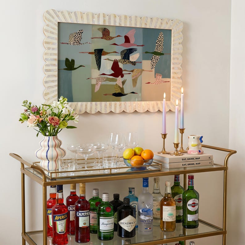 May include: A gold metal bar cart with two glass shelves holding various liquor bottles, glassware, a bowl of citrus, and three taper candles. A scalloped frame surrounds a print of flying birds, and a vase with flowers sits on top.