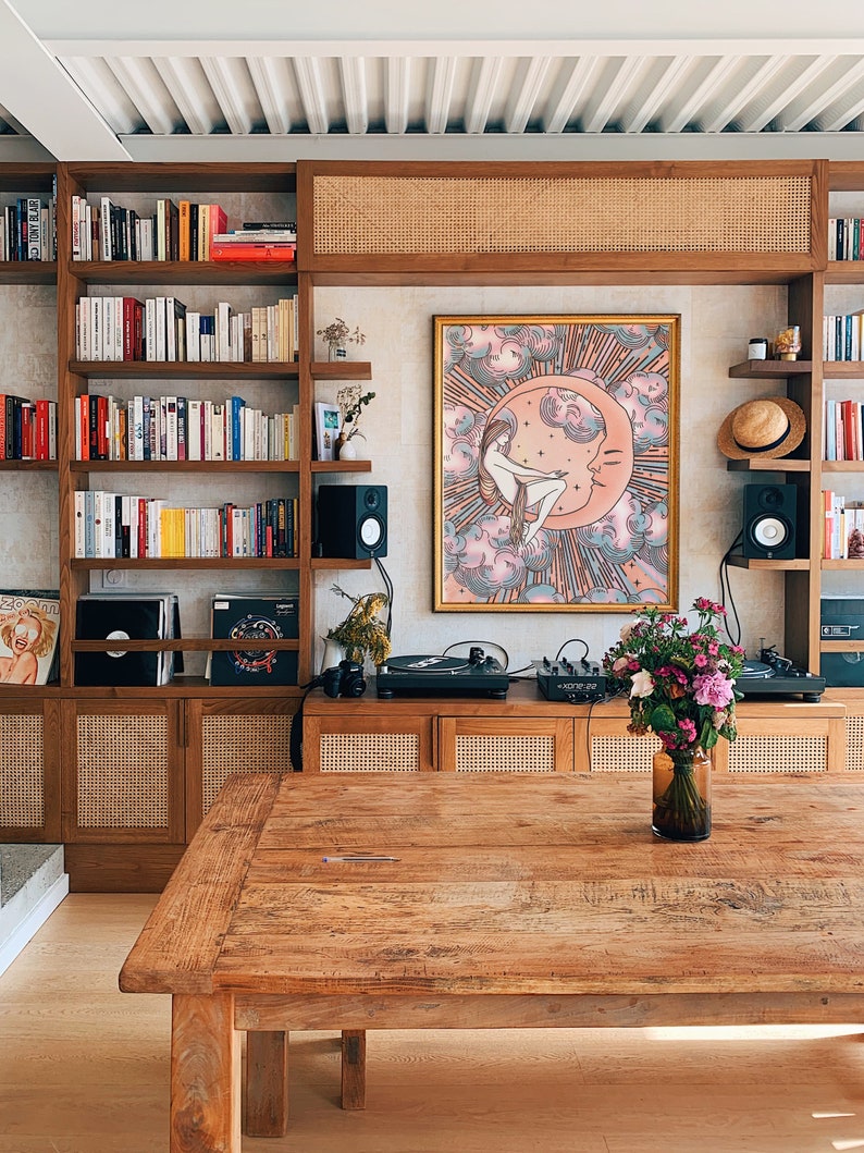 May include: A wooden table with a light wood finish sits in a room with a bookshelf, a record player, and a framed print of a woman sitting on a crescent moon. The print has a pink, blue, and gold color scheme.
