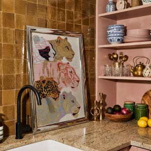 May include: A rectangular framed print featuring four stacked big cat heads in shades of tan, pink, orange, and blue. The artwork is displayed in a silver frame, positioned on a granite countertop next to a black tap and kitchen shelving.