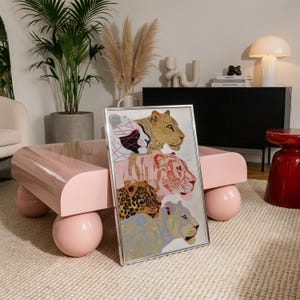 May include: A rectangular framed wall art piece featuring a vertical stack of four stylised big cats. The top cat is gold, the second is pink with white stripes, the third is a leopard with black spots, and the bottom cat is light blue with gold patterns.
