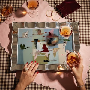 May include: A rectangular serving tray with a mirrored, wavy frame featuring a central illustration of various birds in flight against a segmented blue and green background. The tray is surrounded by three glasses of amber liquid and a pearl necklace.