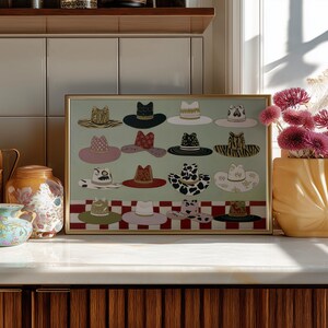 May include: A framed print featuring a collection of illustrated hats in various styles and patterns. The hats are arranged on a light green background with a red and white checkered border. The artwork is displayed on a white countertop.