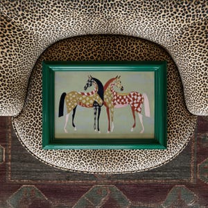 May include: A framed artwork featuring two stylized horses with patterned coats in shades of gold, red, and black. The green frame contrasts with the animal print chair it rests on. The artwork is placed on a patterned rug.