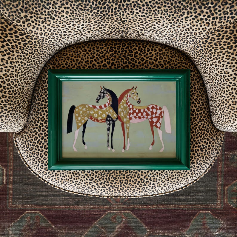 May include: A framed artwork featuring two stylized horses with patterned coats in shades of gold, red, and black. The green frame contrasts with the animal print chair it rests on. The artwork is placed on a patterned rug.