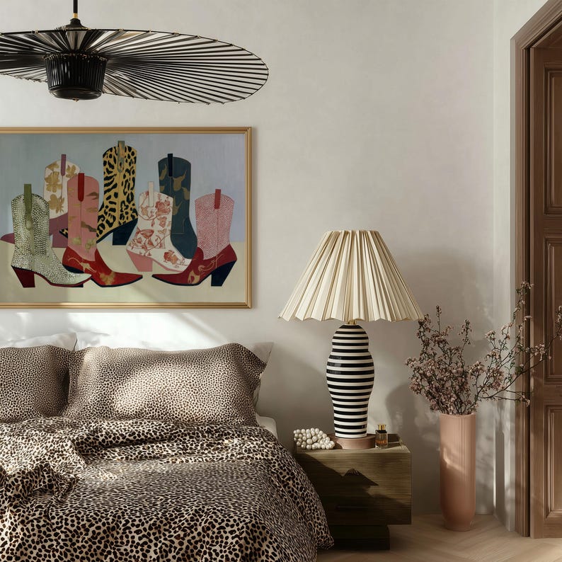 May include: A bedroom scene featuring a bed with leopard print bedding. A framed artwork of cowboy boots hangs above the bed. A lamp with a striped base and pleated shade sits on a wooden nightstand. A tall pink vase holds dried flowers.