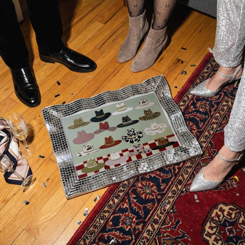 May include: A decorative mirrored tray featuring a collection of illustrated hats in various colors and patterns. The tray is surrounded by a mosaic border and rests on a wooden floor. The hats include designs such as a black fedora, a pink hat with a bow, and a cow-print hat.
