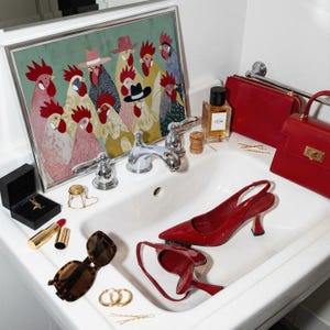 K&ouml;nnte beinhalten: Ein Waschbecken mit Modeaccessoires und Dekoration. Ein gerahmtes Kunstwerk mit H&auml;hnen, rote Abs&auml;tze, Handtaschen, Sonnenbrillen, Schmuck und Parf&uuml;mflaschen sind auf dem wei&szlig;en Porzellan angeordnet. Ein silberner Wasserhahn befindet sich in der Mitte.