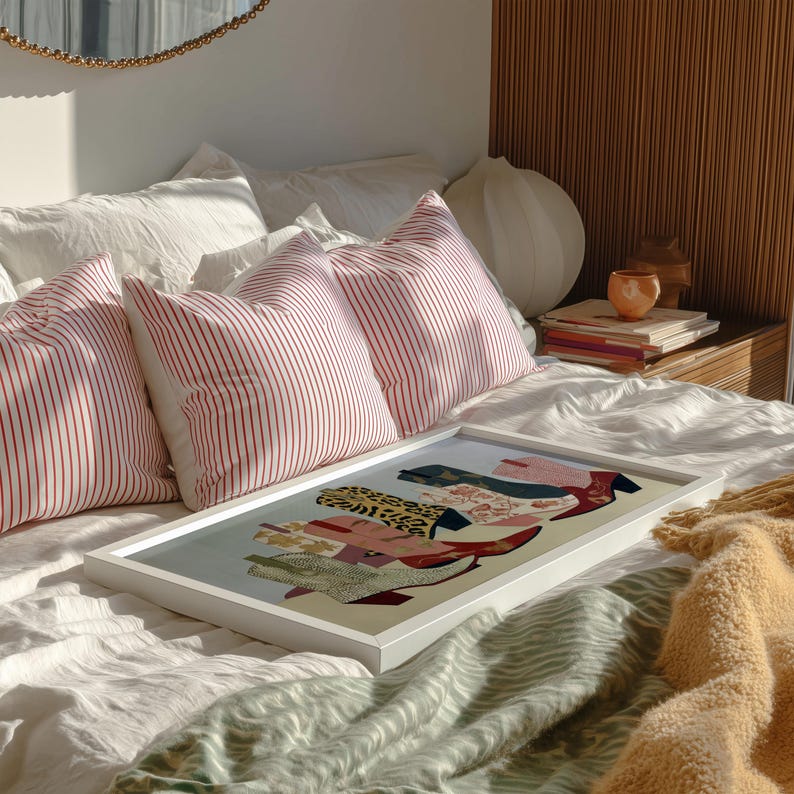 May include: A framed artwork featuring a collage of colorful boots and shoes rests on a bed with white linens. Red and white striped pillows and a yellow blanket add color. A round mirror and a wooden nightstand are in the background.