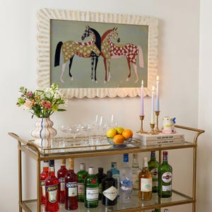 May include: A gold bar cart with a variety of liquor bottles, glassware, and a vase of flowers. Above, a framed painting of two horses with patterned coats hangs on the wall. Candles and books are also on the cart.