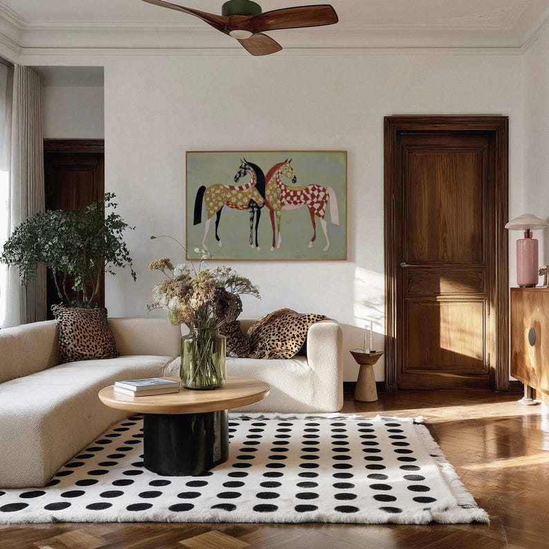 May include: A living room scene featuring a cream-colored sofa with leopard print pillows, a round wooden coffee table, and a black and white polka dot rug. A painting of two horses hangs above the sofa.