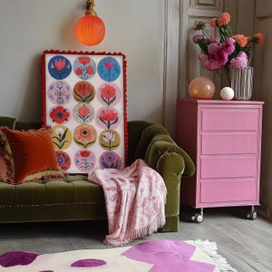 May include: A living room scene featuring a green velvet sofa with orange pillows, a pink dresser, and a framed floral art piece. A pink throw blanket and a patterned rug add color. A decorative orange light fixture is mounted on the wall.